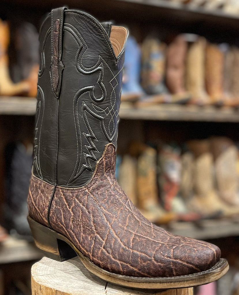 Brown and black cowboy boot on a wooden block with a store shelf of boots in the background