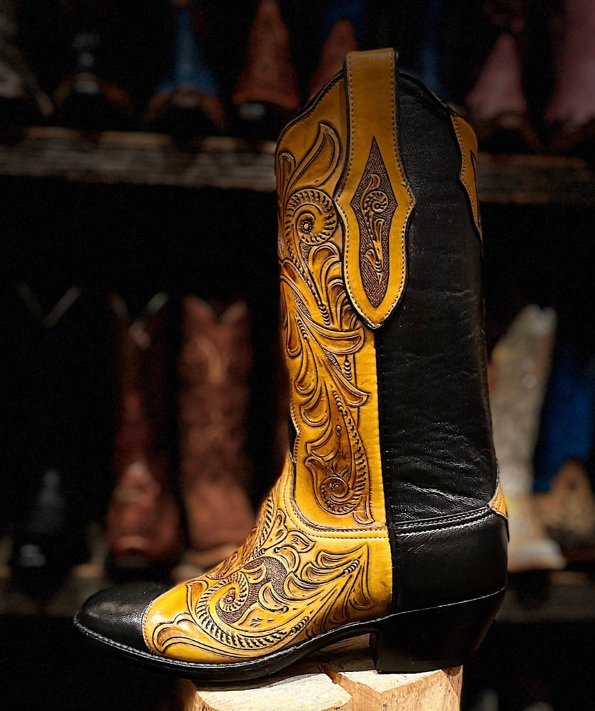 Cowboy boot with intricate yellow leather design on a wooden block, blurred background of boots.