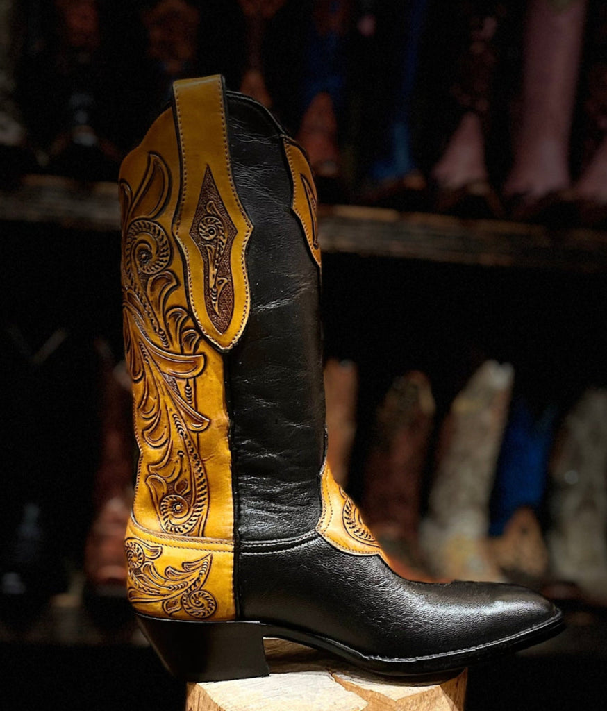 Black cowboy boot with yellow tooled leather on a wooden block against a blurred background