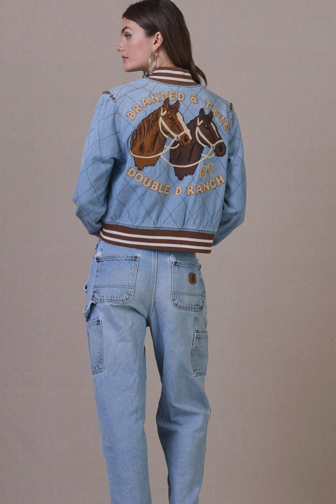 Woman wearing a blue denim jacket with horse designs and text, standing against a plain background.