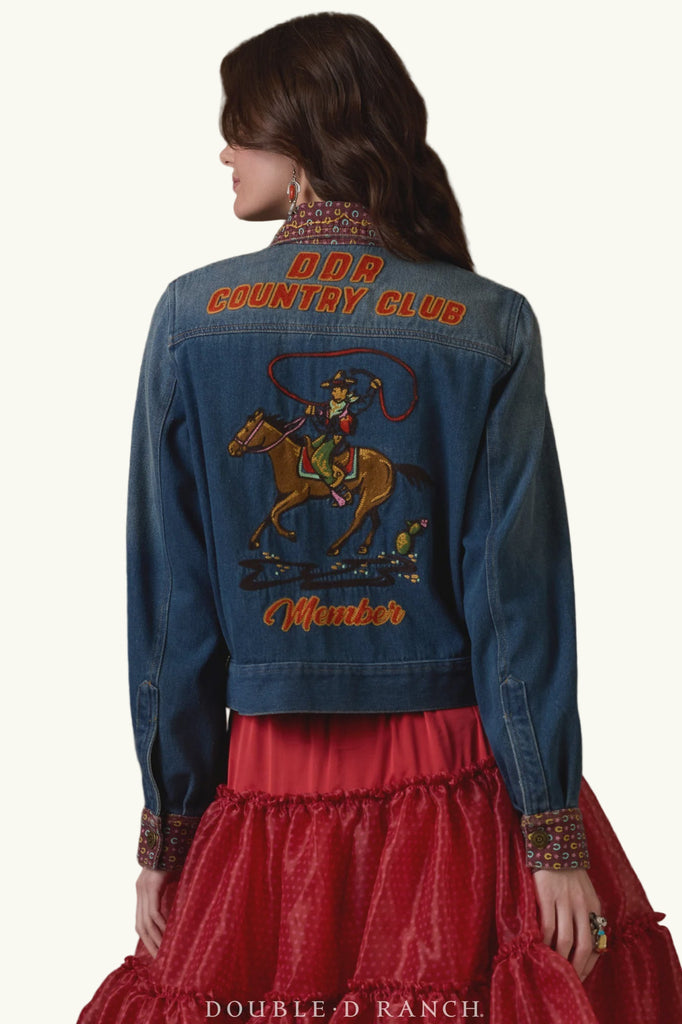 Woman wearing a blue denim jacket with embroidered designs and text, standing against a brown background.