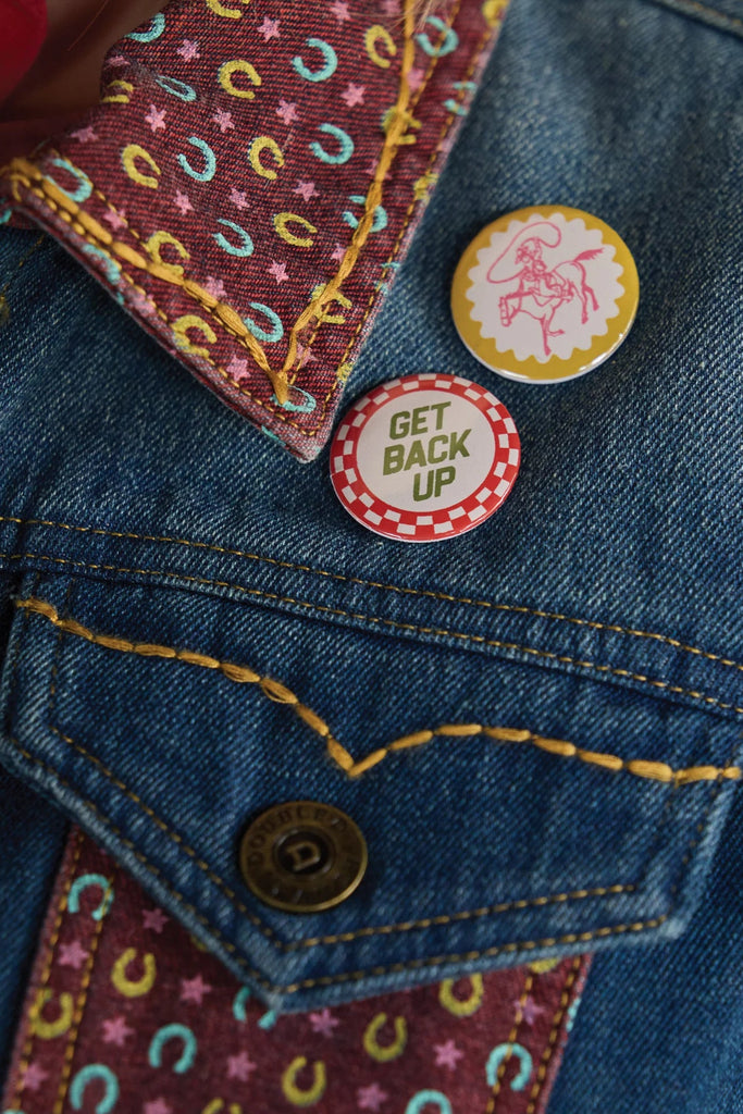 Close-up of a denim jacket with colorful patterns and pins, including one that says 'Get Back Up'.