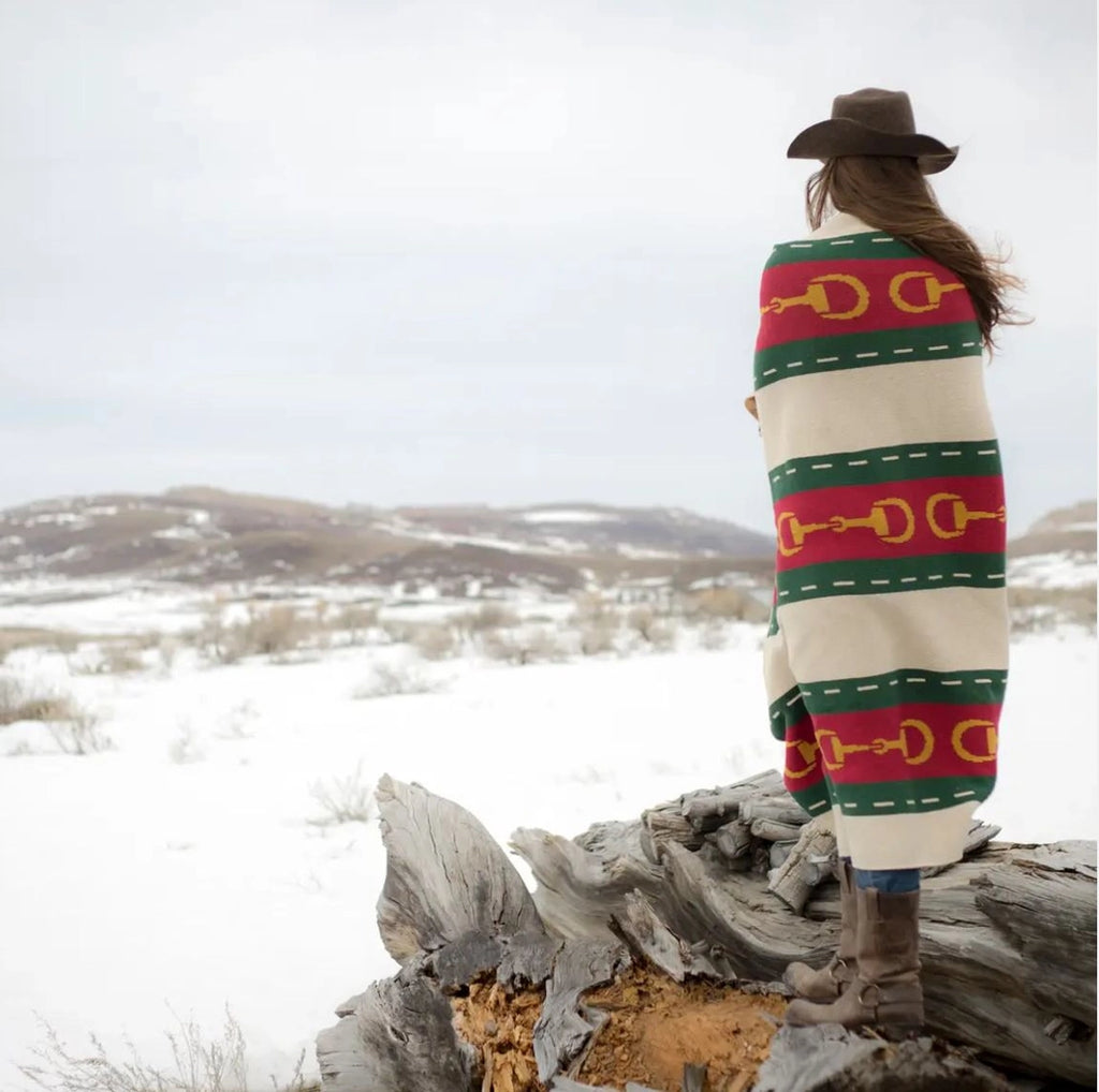 Person wrapped in a colorful blanket with horse shoe pattern, standing on a log in a snowy landscape.