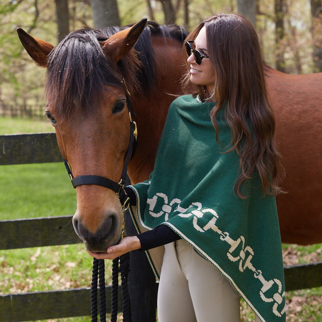A woman wearing a green Equestrian Snaffle Bit Poncho with a chain pattern, standing beside a brown horse.