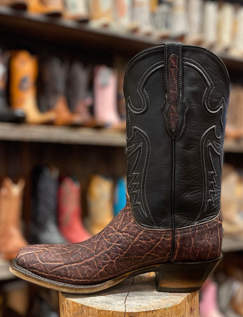 Brown and black exotic cowboy boot on a wooden block with a blurred boot store background