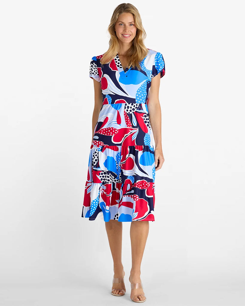 A woman modeling a short-sleeve midi dress with a vibrant abstract print in red, blue, white, and navy, set against a plain white studio background.