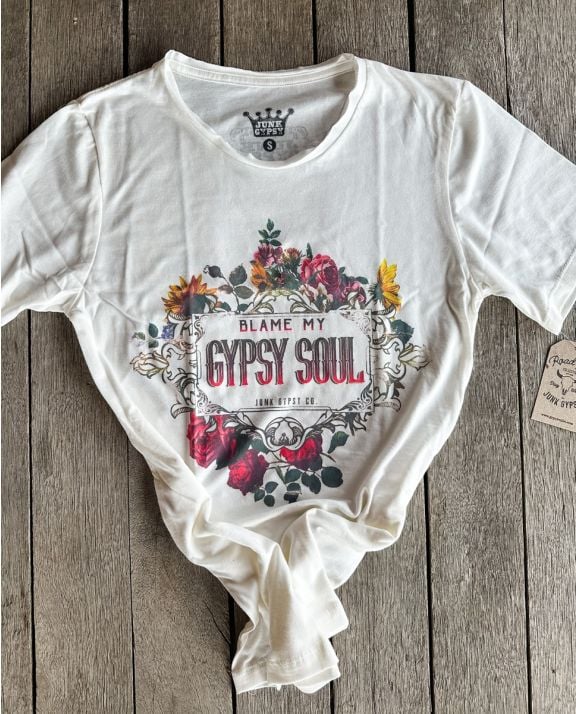A white short-sleeve T-shirt with a colorful graphic print on the front featuring flowers and the text 'BLAME MY GYPSY SOUL'. The shirt is styled with lots of color and is laid flat on a rustic wooden plank background. 100% cotton