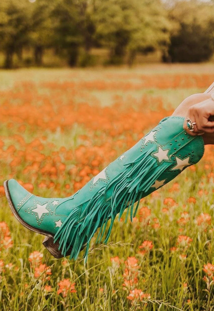 Teal cowboy boots with star patterns and fringes worn by a person in a field of orange flowers.