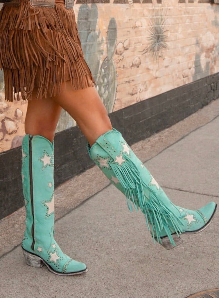 Teal cowboy boots with fringe and star patterns worn by a person on a sidewalk.