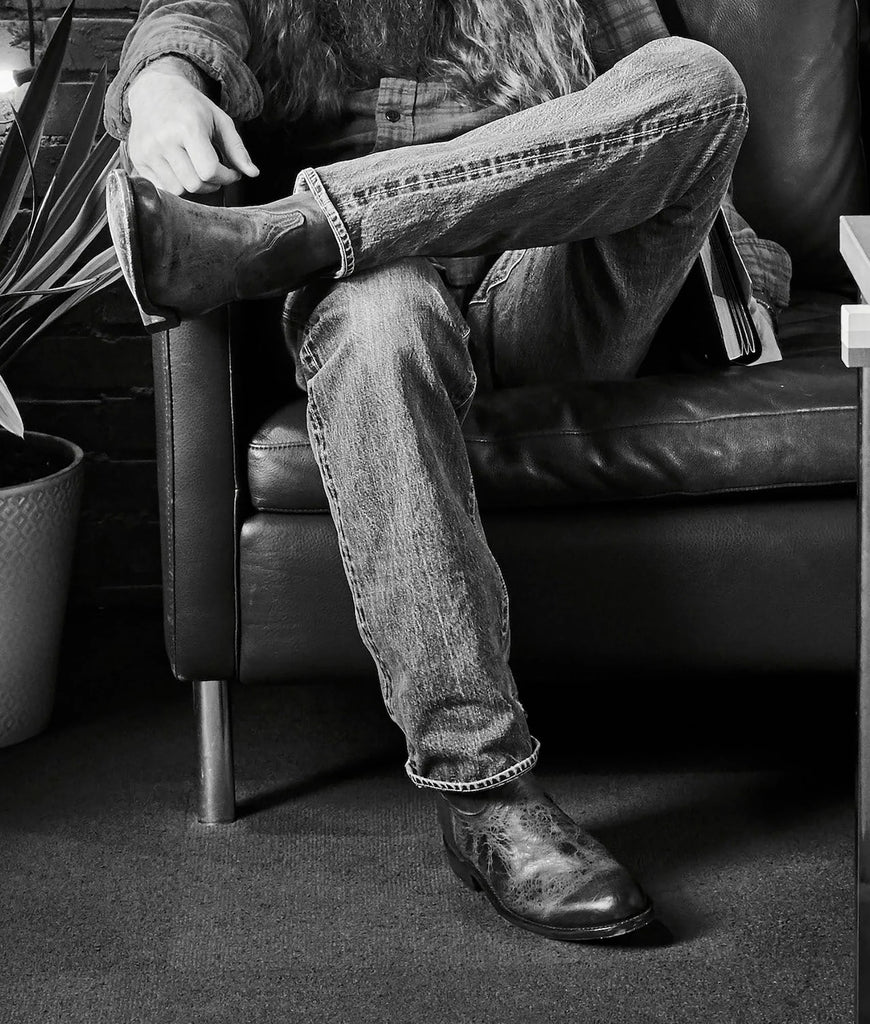 Cowboy sitting on a leather couch wearing jeans and shoes, with a plant and table in the background.