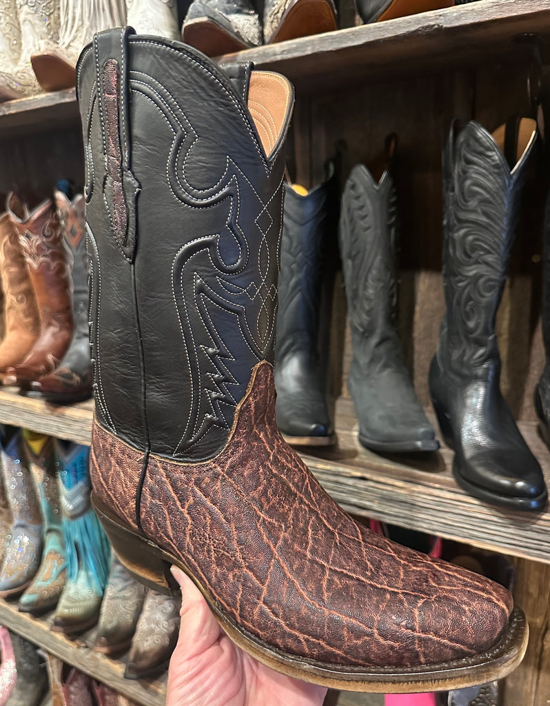 Brown elephant leather cowboy boot with intricate design held in front of a display of other boots.
