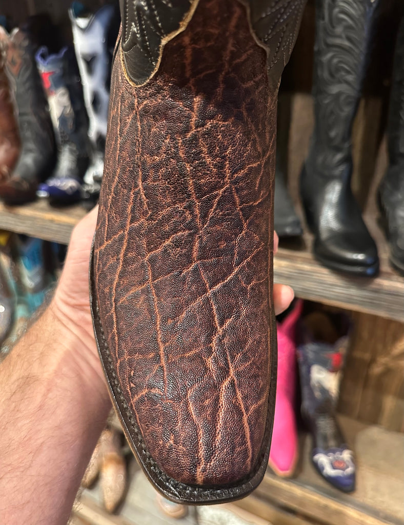 Close-up of a brown leather boot sole held by a hand with a shoe rack in the background.