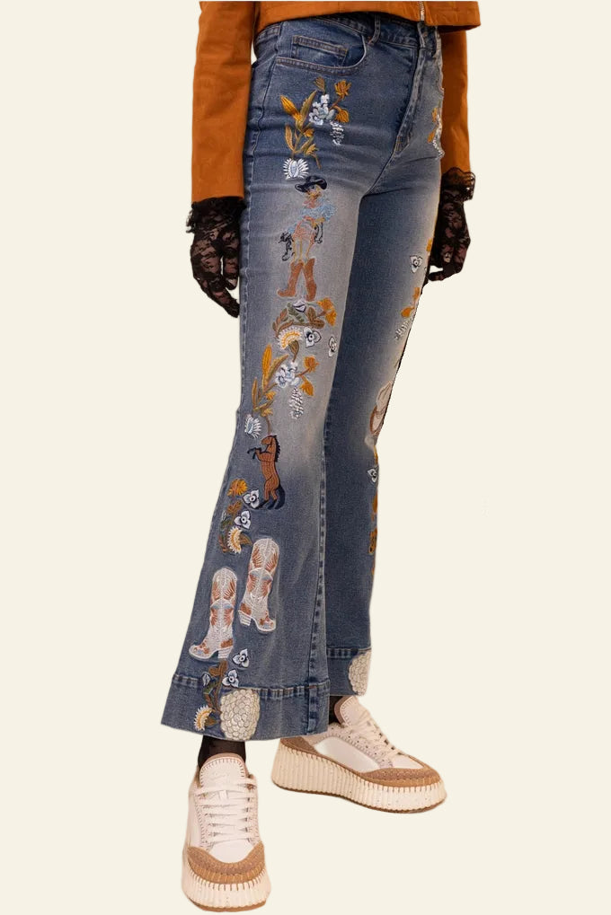 Person wearing blue jeans with embroidered designs, holding a black handbag on a beige background