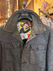 A dark gray wool-blend button-front jacket hanging on a black hanger with a vintage comic-style collar label, photographed inside a retail store.