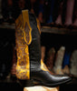 Black cowboy boot with yellow tooled leather on a wooden block against a blurred background