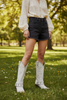 A person standing outdoors on grass wearing dark denim shorts, a cream blouse, and off-white western-style boots with decorative trim.