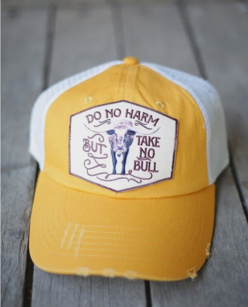 A distressed yellow and white cap hat with a front patch that reads 'DO NO HARM BUT TAKE NO BULL' and features a cow illustration, shown on a gray wooden surface.