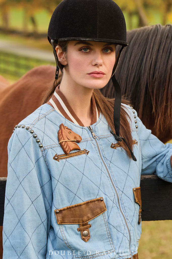 Woman in equestrian attire with a horse emblem, standing next to a horse in an outdoor setting.