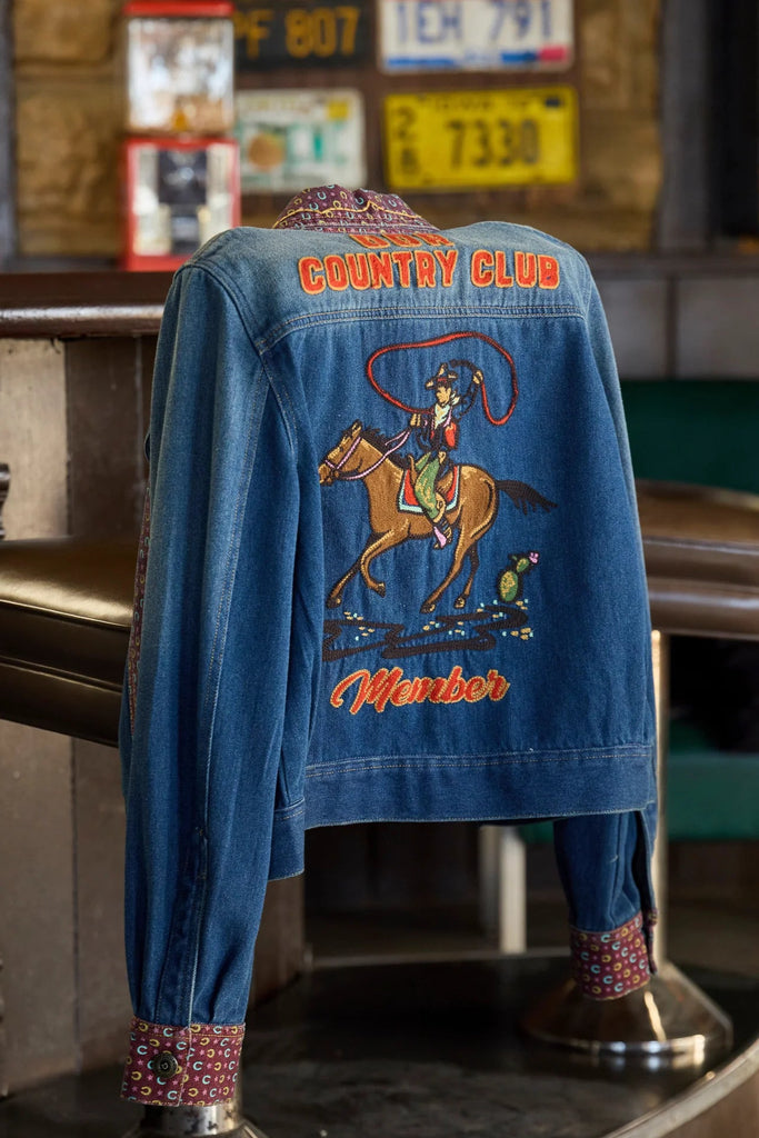 Blue denim jacket with cowboy graphics and text on a stool in a bar setting.