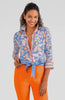 Woman wearing a blue floral blouse with a tie and orange pants on a gray background