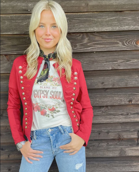A blonde woman wearing a red military-style jacket with gold buttons over a white graphic t-shirt and blue jeans, standing against a rustic wooden plank wall.