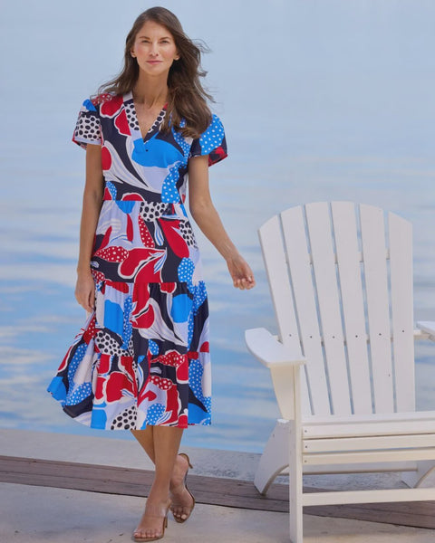 Woman in a colorful dress standing by a white chair on a dock with water in the background