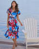Woman in a colorful dress standing by a white chair on a dock with water in the background
