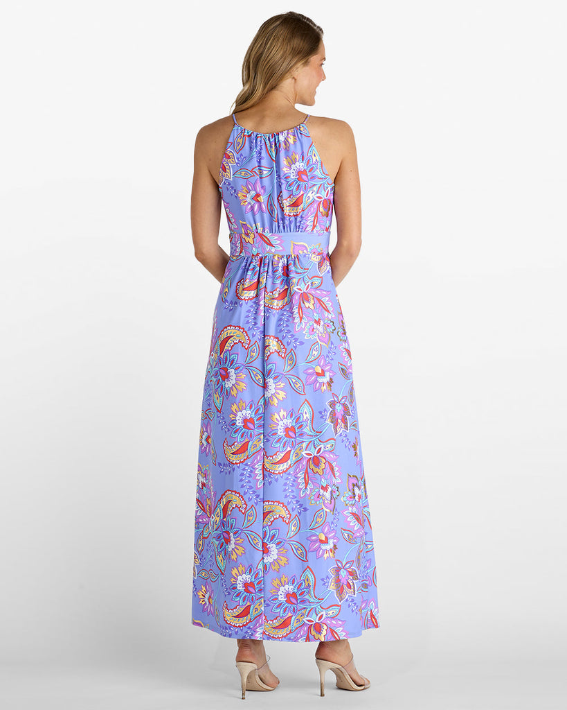 A woman shown from the back wearing a sleeveless halter-neck maxi dress with a colorful paisley and floral print on a light purple background, paired with beige high heels, against a plain white studio background.