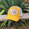 A yellow and white cap hat with a distressed brim, resting on a wooden fence in front of green plants. The front patch features a cartoon bull and the text 'DO NO HARM BUT TAKE NO BULL'.