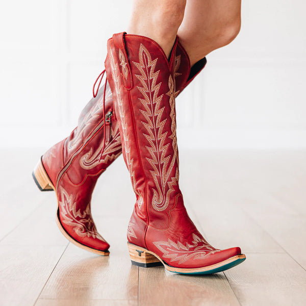Red cowboy boots with intricate white embroidery on a light wooden floor.