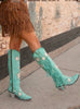 Teal cowboy boots with fringe and star patterns worn by a person on a sidewalk.
