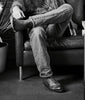 Cowboy sitting on a leather couch wearing jeans and shoes, with a plant and table in the background.