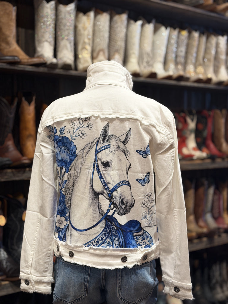 A white denim jacket with a large blue and white graphic print of a horse on the back, displayed on a mannequin in a store with cowboy boots on shelves in the background.