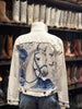 A white denim jacket with a large blue and white graphic print of a horse on the back, displayed on a mannequin in a store with cowboy boots on shelves in the background.