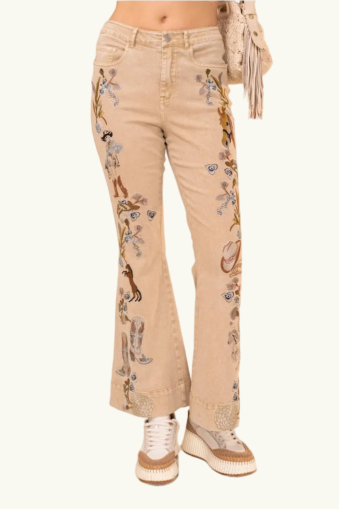 Beige jeans with floral horse and cowgirl embroidery worn by a person, with an American flag in the background.